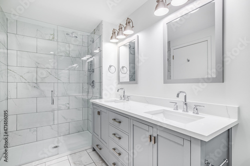 An elegant, remodeled bathroom with a grey vanity and bronze hardware. The shower has a large shower head and marble tiles and glass wall line the sides.