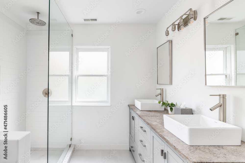 An elegant, renovated bathroom with white sinks, grey vanity, granite countertop, and bronze hardware, faucets and light. The shower is lined with white tiles. An elegant, renovated bathroom with white sinks, grey vanity, granite countertop, and bronze hardware, faucets and light. The shower is lined with white tiles.
