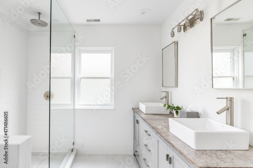 An elegant, renovated bathroom with white sinks, grey vanity, granite countertop, and bronze hardware, faucets and light. The shower is lined with white tiles.