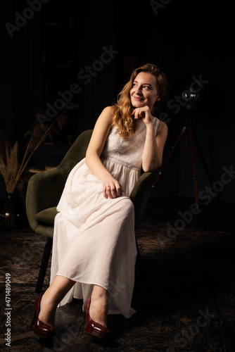 A young woman in a dress with long hair smiles and looks to the side while sitting in a chair in a dark room.