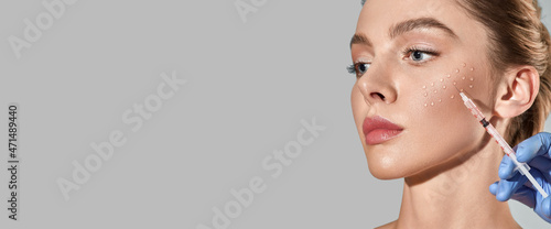 Biorevitalization procedure. Pretty woman while beauty injections with hyaluronic acid for smoothing of mimic wrinkles around eyes, over grey background with space for text photo