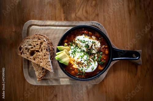 Top view. Shakshuka with two poached eggs and avocado in a black pan and bread aside on a wooden board. Middle eastern traditional dish. Flat lay photo