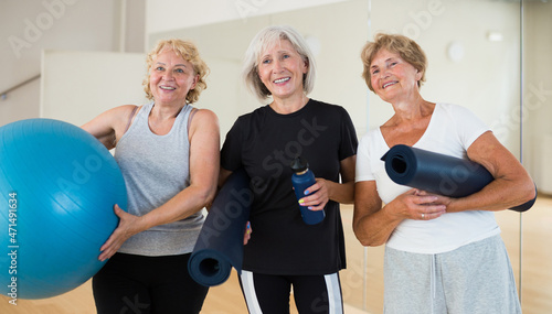 Group photo of smiling mature women standing with fitnessball and rugs inside exercising room.