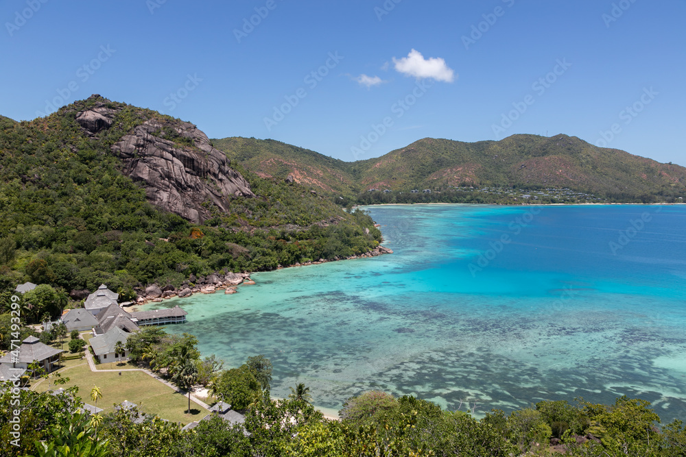 Panoramic view to Anse Petite Cour on Praslin island in the Seychelles from a high viewpoint. Bungalow hotel on a beach with a crystal blue lagoon, endless ocean on a summer day. Tropical landscape.