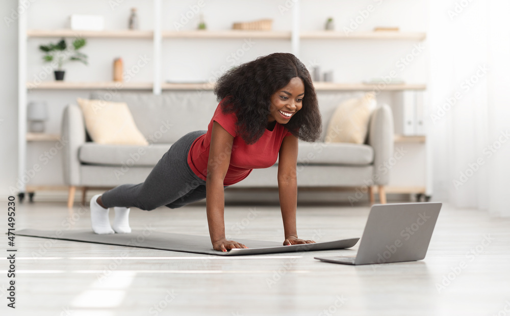 Fototapeta premium Positive african american athletic woman exercising at home, using laptop