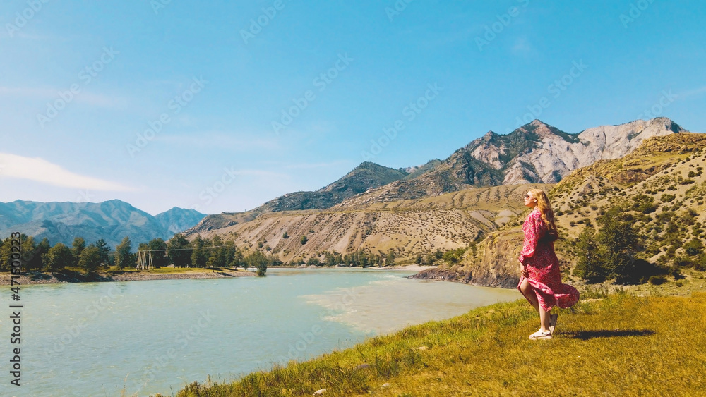 Lady in long red dress overlooks the edge of two rivers with mountains on the background. Drone footage
