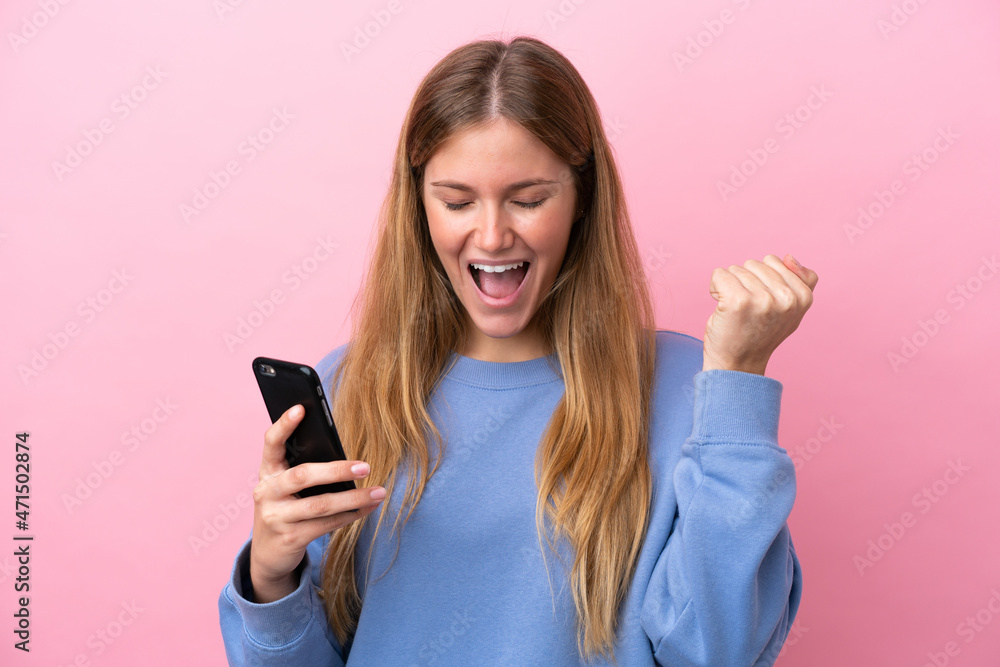 Young blonde woman isolated on pink background using mobile phone and doing victory gesture Young blonde woman isolated on pink background using mobile phone and doing victory gesture