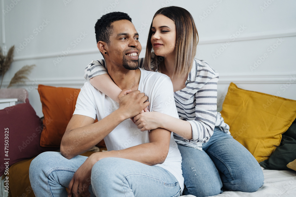 Portrait of happy young African Caucasian couple hug sitting on couch at home, smiling mixed race husband and wife spend time together, embrace relaxing on sofa. Healthy relationships concept Portrait of happy young African Caucasian couple hug sitting on couch at home, smiling mixed race husband and wife spend time together, embrace relaxing on sofa. Healthy relationships concept