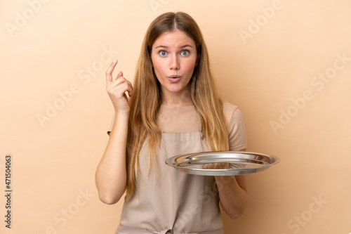Young blonde waitress woman with tray isolated on beige background intending to realizes the solution while lifting a finger up