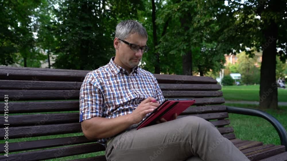 Mature man with grey hair in glasses and colorful shirt sits on bench in park and browses social networks on tablet slow motion Mature man with grey hair in glasses and colorful shirt sits on bench in park and browses social networks on tablet slow motion