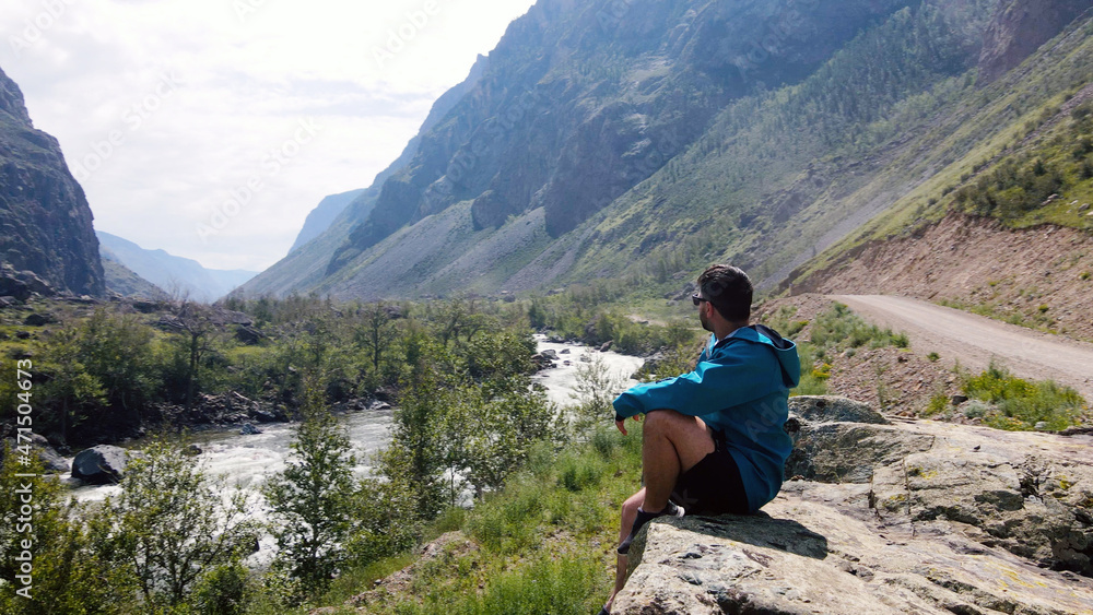 Naklejka premium A man with glasses sits on the edge of a cliff wearing a blue anorak. Katu-Yaryk canyon Chulyshman Valley. Altai