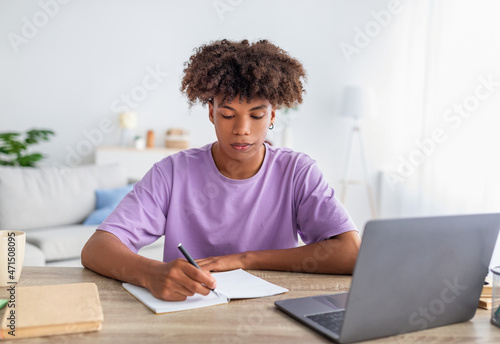 Serious black teen guy using laptop, writing in notebook during online lesson, sitting at desk indoors