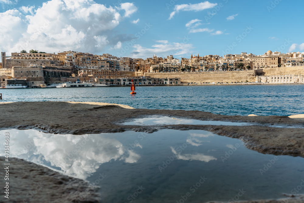 Panoramic view of Valletta,Malta.City skyline from Birgu Vittoriosa harbor.Peaceful cityscape,sunny summer day,clouds reflected in water.Waterfront houses and Upper Barrakka Gardens.Spot for vacation