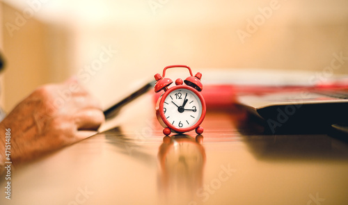 Um pequeno relógio despertador vermelho sobre uma mesa e ao fundo uma senhora usando um celular smartphone durante o horário do seu trabalho em casa. Distração, procrastinação, improdutividade.