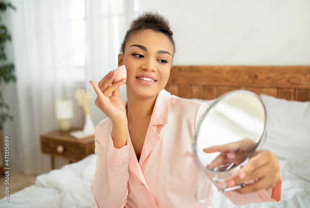 Pretty african american lady applying cosmetic foundation with applicator, holding mirror, sitting on bed in bedroom Pretty african american lady applying cosmetic foundation with applicator, holding mirror, sitting on bed in bedroom