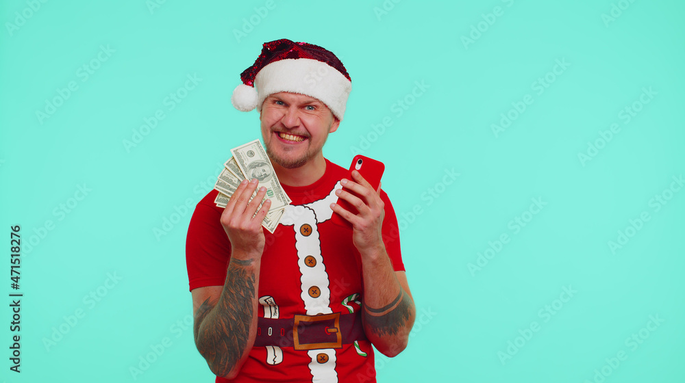 Obraz premium Man in Christmas red t-shirt and hat looking mobile phone display sincerely rejoicing win, receiving money dollar cash banknotes, success lottery luck. Guy isolated on blue background. Happy New Year