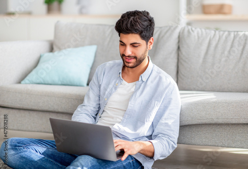 Young arab freelancer sitting with laptop on floor and leaning on sofa while working on computer at home interior