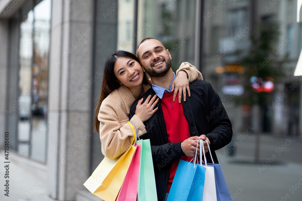 Attractive intrracial couple embracing and smiling at camera, going shopping, posing with gift bags near supermarket Attractive intrracial couple embracing and smiling at camera, going shopping, posing with gift bags near supermarket