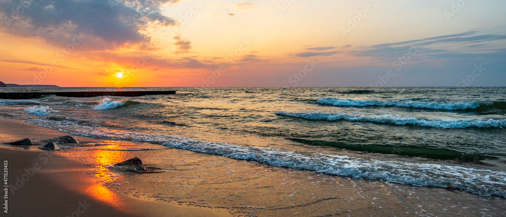 Sunset in the wavy sea with a wooden breakwater and stones on the sandy shore, beautifully colored sky