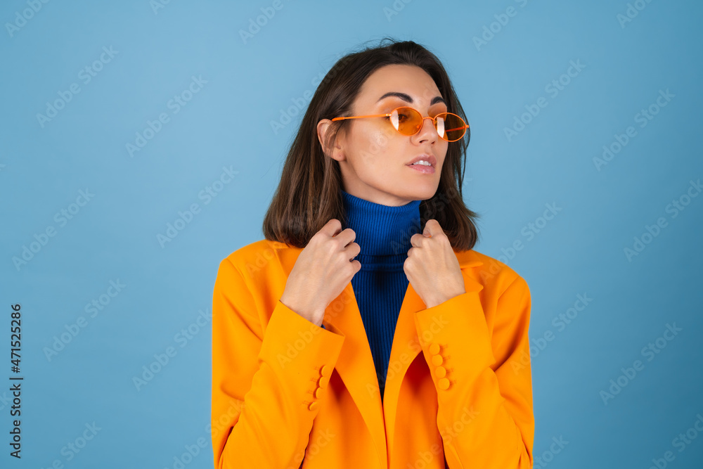 Fashionable young woman in a knee-length and oversized jacket on a blue background in stylish bright orange glasses posing Fashionable young woman in a knee-length and oversized jacket on a blue background in stylish bright orange glasses posing
