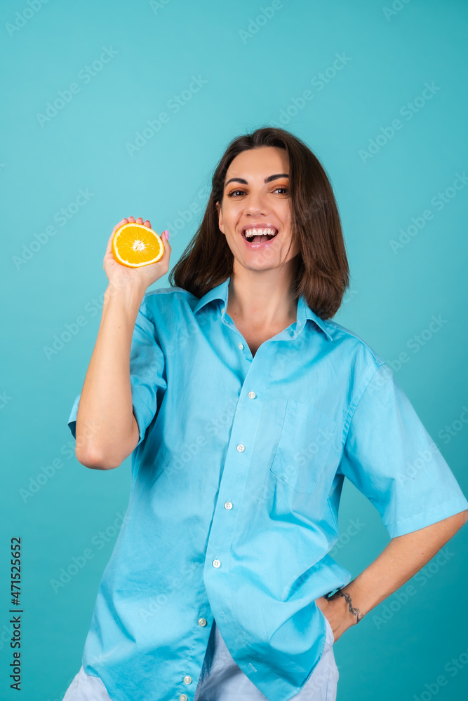 Young woman in a blue shirt on the background holds an orange, poses cheerfully, in high spirits, laughs smiles