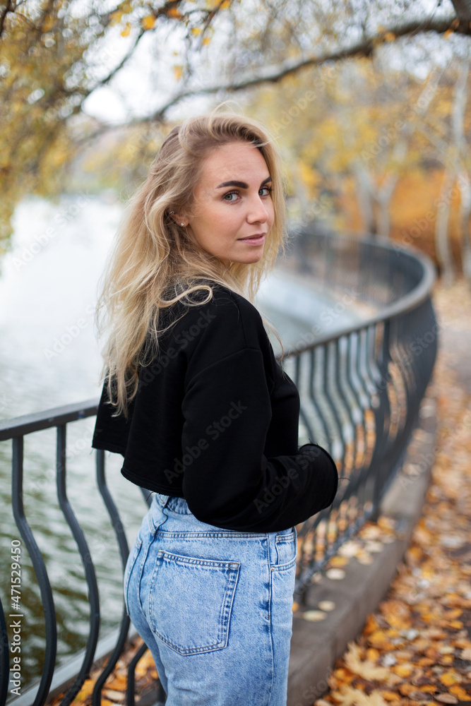 beautiful caucasian blonde female in black sweater jeans walking near a pond in the autumn park beautiful caucasian blonde female in black sweater jeans walking near a pond in the autumn park