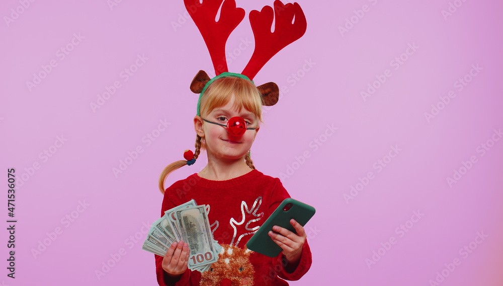 Girl in red Christmas deer antlers looking mobile phone display sincerely rejoicing win, receiving money dollar cash banknotes, success lottery luck. Kid isolated on violet background. Happy New Year