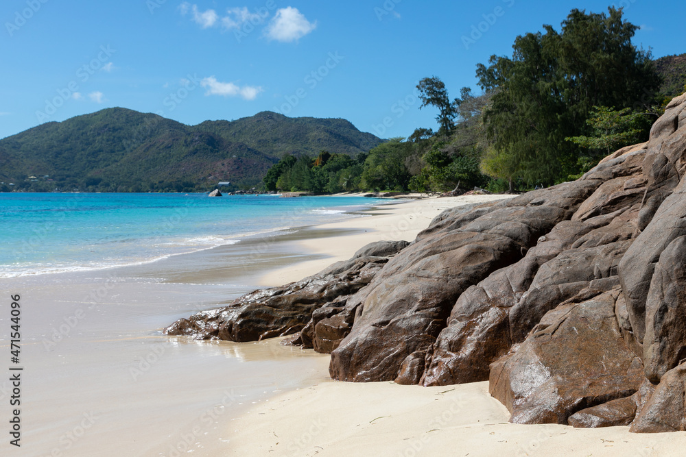 A solid brown granite formations grow from white sands on Anse Boudin beach in the Seychelles. Turquoise crystal waters of the ocean wash the smooth coastline. Behind the hills and tropical jungles.