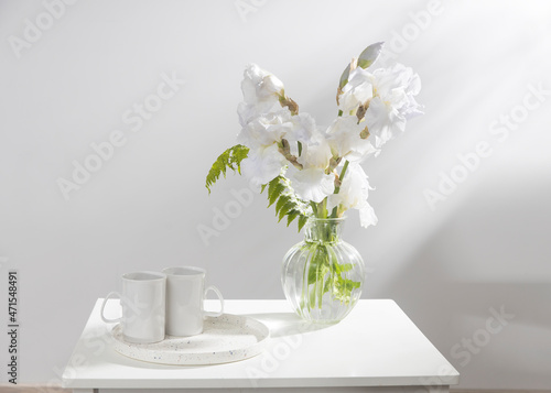 A bouquet of three white irises and a fern in a transparent vase on the table. Two ceramic tea cup in the tray. Breakfast