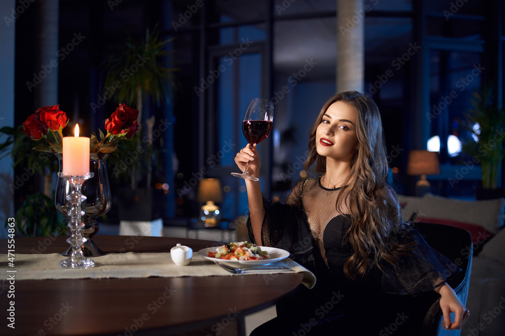 Gorgeous woman in stylish black dress sitting at table with fresh vegetable salad and glass of red wine. Candles and flowers as decoration. Romantic mood. Gorgeous woman in stylish black dress sitting at table with fresh vegetable salad and glass of red wine. Candles and flowers as decoration. Romantic mood.