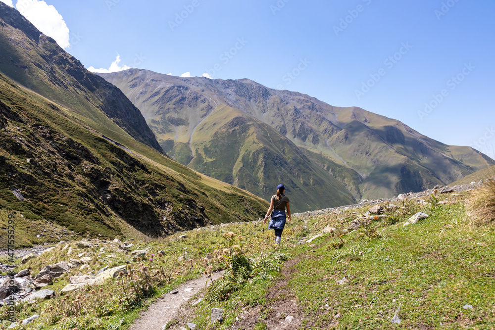 A female backpacker on a hiking trail with a panoramic view on the green hills and sharp ridges of the mountain peaks in the Greater Caucasus Mountain Range in Georgia, Kazbegi Region. Wanderlust.