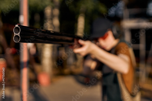 woman in forest standing in the attitude of aiming and looking through the sight of automatic rifle, going to shoot. caucasian lady in protective eyeglasses, cap and headphones. focus on rifle