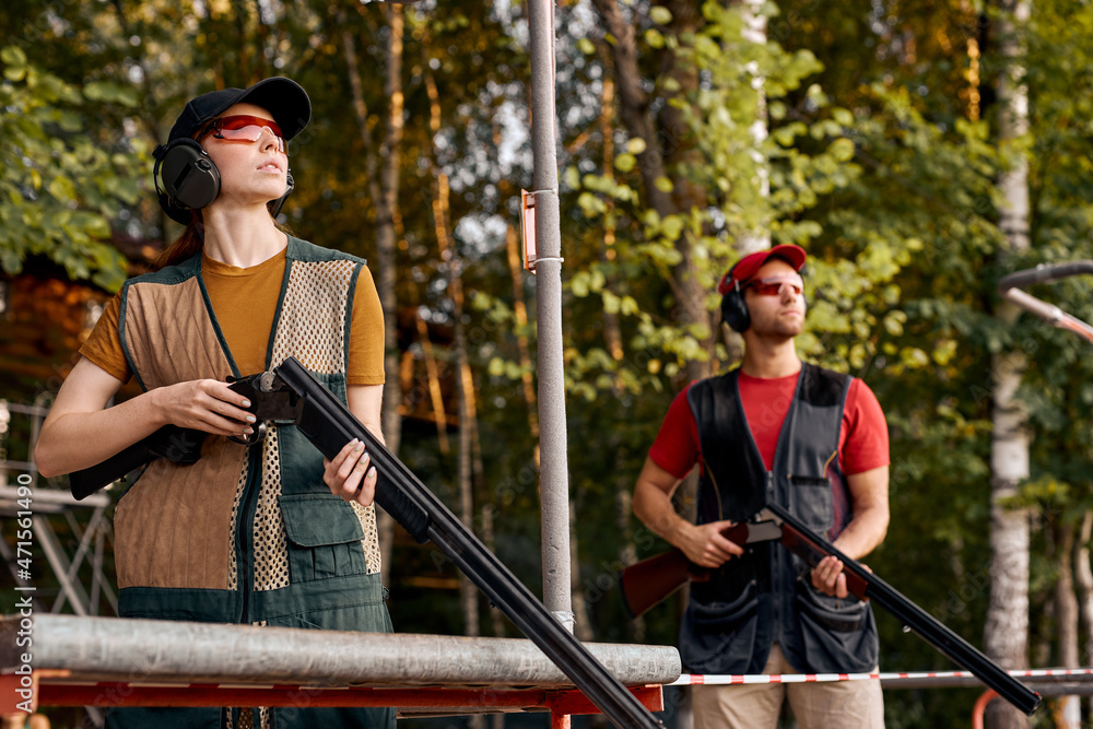 © Roman - Side View On Male And Female In Outdoor Range, Looking At Side At Target, After Shooting, Concentrated on Training, At Summer Season, Wearing Protective Equipment Uniform, Goggles And Headset