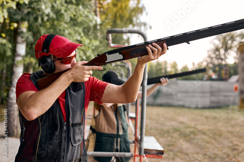 After getting shooting instructions, young caucasian man concentrated at aiming rifle at side, fires at target in outdoor range, wearing goggles, cap and headset equipment outfit. Side view