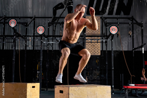 Fotografia Young caucasian shirtless male is jumping on cross fit wooden box, at modern sportive gym