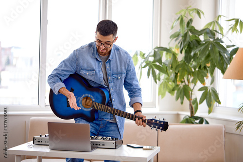 Man in denim jacket playing acoustic guitar  using laptop  performing music in studio  standing behind desk with piano  window in the background. Smiling caucasian male preparing for concert