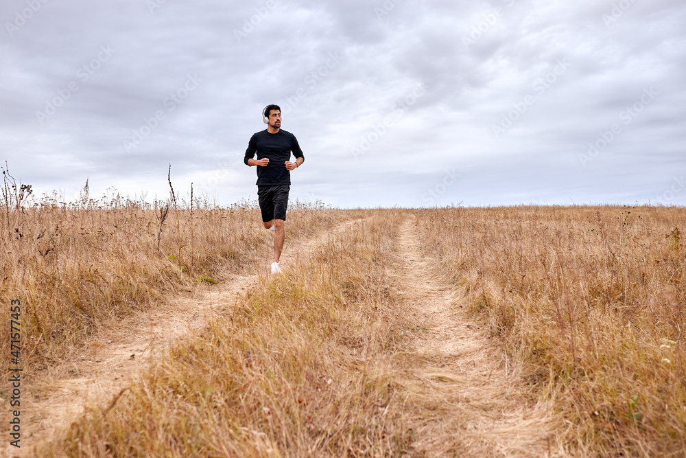 Man running,jogging, sprinting in mountains. Fit male fitness runner during outdoor workout. Young athlete caucasian man in black sportswear tracksuit and headphones earphone listening to music.