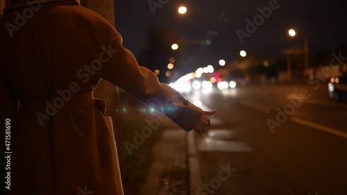 a girl in a coat stands near the road at night and raises her hand to stop cars.