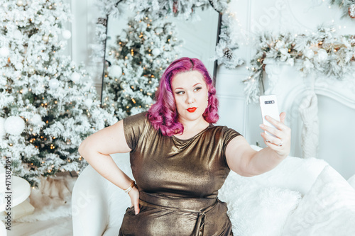 Beautiful young Caucasian plus size model woman in long golden olive color dress taking selfie picture photo portrait in holiday winter decoration at home .