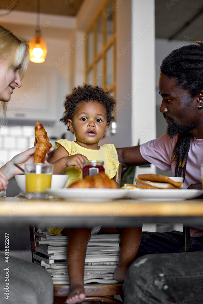 Happy young family with black cute daughter having breakfast together at table in modern bright cozy kitchen, man woman and kid eating healthy food at home, laughing having talk. friendly family Happy young family with black cute daughter having breakfast together at table in modern bright cozy kitchen, man woman and kid eating healthy food at home, laughing having talk. friendly family