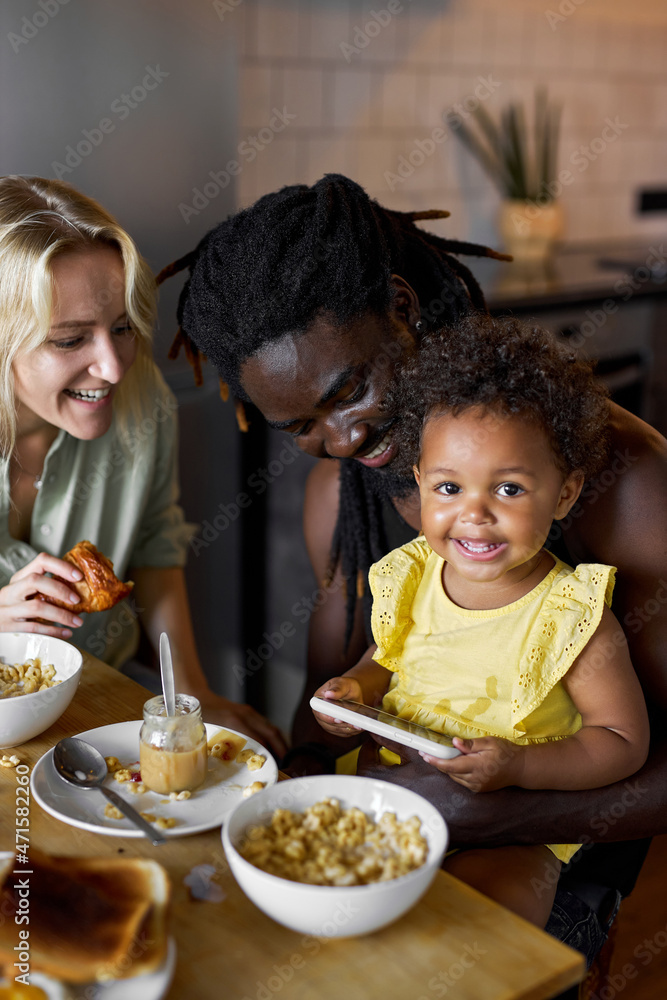 happy child girl watching video cartoons on smartphone while having meal with parents, free time, leisure in the morning. pleasant caucasian and black parents sit with child enjoying time together