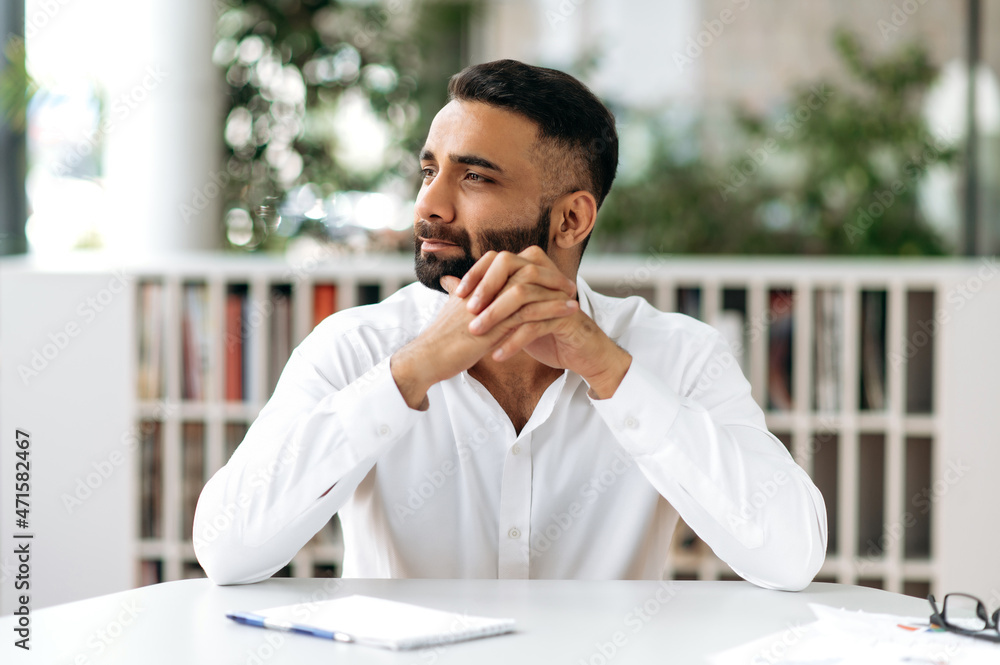 Naklejka premium Concentrated Indian man, businessman, manager, wearing white shirt, sitting at the table in his office, looking pensively to the side, thinking about a new project or dreaming of a vacation