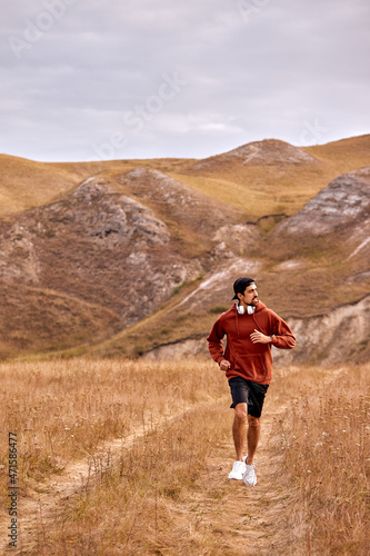 Sportive athlete adult man in red hoodie is running jogging, listen to music, enjoy sport. wonderful landscape, mountains in the background. man is in action, strong and fit. cardio training
