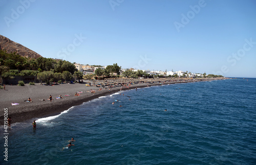 Fototapeta Naklejka Na Ścianę i Meble -  People sunbathing and swimming at black beach of Kamari in Santorini, Greece. Beach is covered with fine black sand, and drops off sharply into the water.