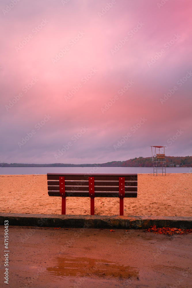 Desolate beach park with an empty bench. Moody seascape with the view of pink clouds, island, lifeguard watchtower, bench, sand, and reflections on the puddle.
