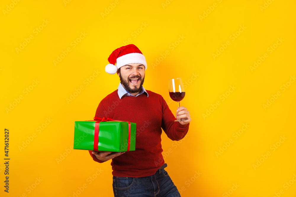 young latin man holding a glass of wine with santa hat and christmas presents on a yellow background in Mexico Latin America young latin man holding a glass of wine with santa hat and christmas presents on a yellow background in Mexico Latin America
