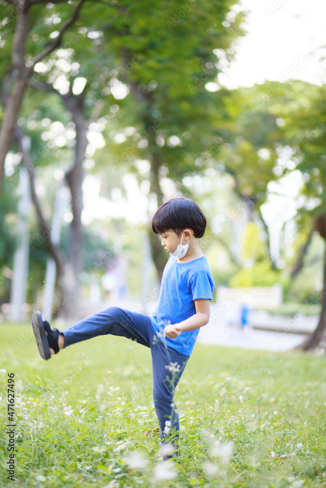 A cute boy wearing a blue shirt, long black pants and a white mask sits in a lush green garden in the spring against a backdrop of green trees. portrait shot. A cute boy wearing a blue shirt, long black pants and a white mask sits in a lush green garden in the spring against a backdrop of green trees. portrait shot.