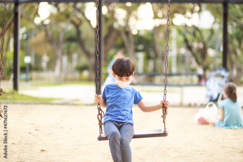 A cute Asian boy wearing a mask is playing on a swing in the playground during the daytime in summer. Outdoor activities. Play Makes Ideas Believe External education. portrait A cute Asian boy wearing a mask is playing on a swing in the playground during the daytime in summer. Outdoor activities. Play Makes Ideas Believe External education. portrait