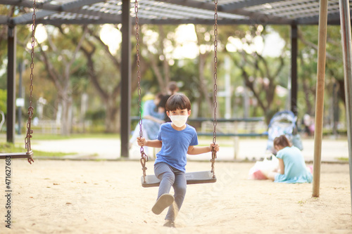 A cute Asian boy wearing a mask is playing on a swing in the playground during the daytime in summer. Outdoor activities. Play Makes Ideas Believe External education. portrait © Tanawit