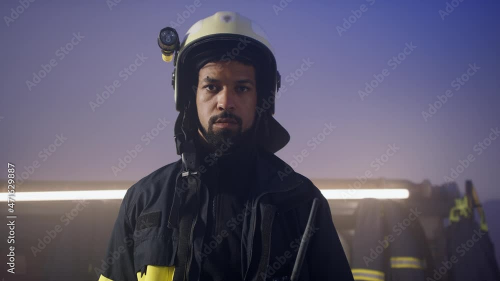 Young African-American firefighter putting on helmet and looking at camera in fire station at night. Young African-American firefighter putting on helmet and looking at camera in fire station at night.